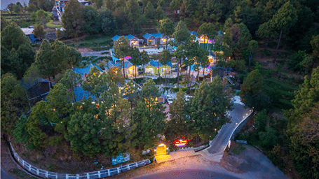 Aerial high-angle shot of DLS Nature Trinket Resort, Dalhousie, showcasing cottages nestled among dense pine trees on a hillside, with an entrance gate leading to a curved paved driveway off a winding mountain road.