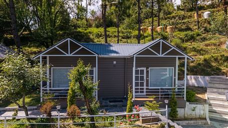Charming hillside cottage with a twin-gabled roof, large front windows, and a landscaped garden surrounded by trees and greenery - DLS Nature Trinket Resort, Dalhousie.