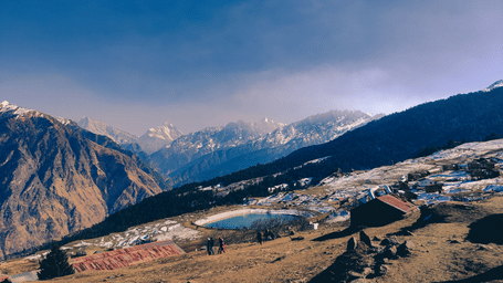 Panoramic view of Auli's mountain landscape with winding trails, snow patches, and distant Himalayan peaks beneath a soft blue sky