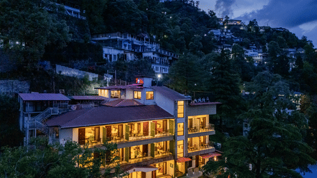 Exterior of Perfectstayz Premium Seven Oaks, Mussoorie, with lit windows and red roofs, stands on a wooded hillside under a dark blue sky.