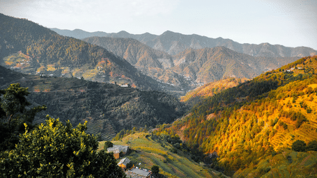 A scenic view of a vast mountain valley with green terraces, where one hillside is cast in deep shadow and the opposite slopes are bathed in warm, golden sunlight.