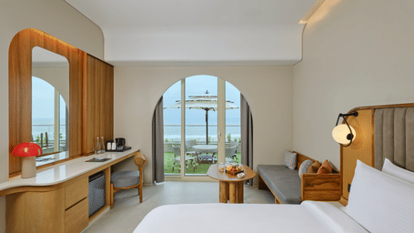 The Premium Room at Swosti Premium Beach Resorts, Puri, featuring a double bed, a television mounted on the wall, modern wooden decor, and large windows displaying a  sea view. 