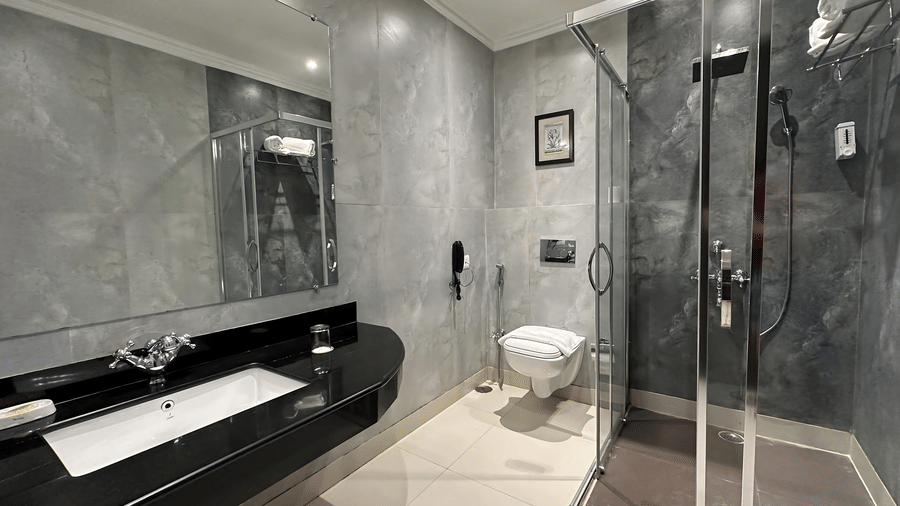 Elegant bathroom in the Deluxe Room at A S Hotels, Khajuraho, featuring a glass walk-in shower, stone-finish wall tiles, and a contemporary black vanity.