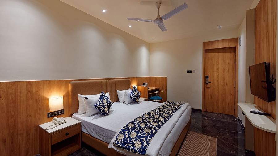 Image of a studio room with a bed with a table on the side of the bed and a television in front Image of a bed with table and chair on the side of the table and a fan on te ceiling at Ankit Hotel, Jabalpur