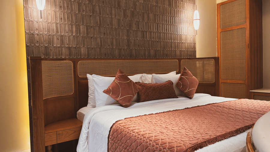 A bed with white linens and a brown patterned runner is flanked by wooden bedside units and wall lamps at Avanya Resorts N Retreat.