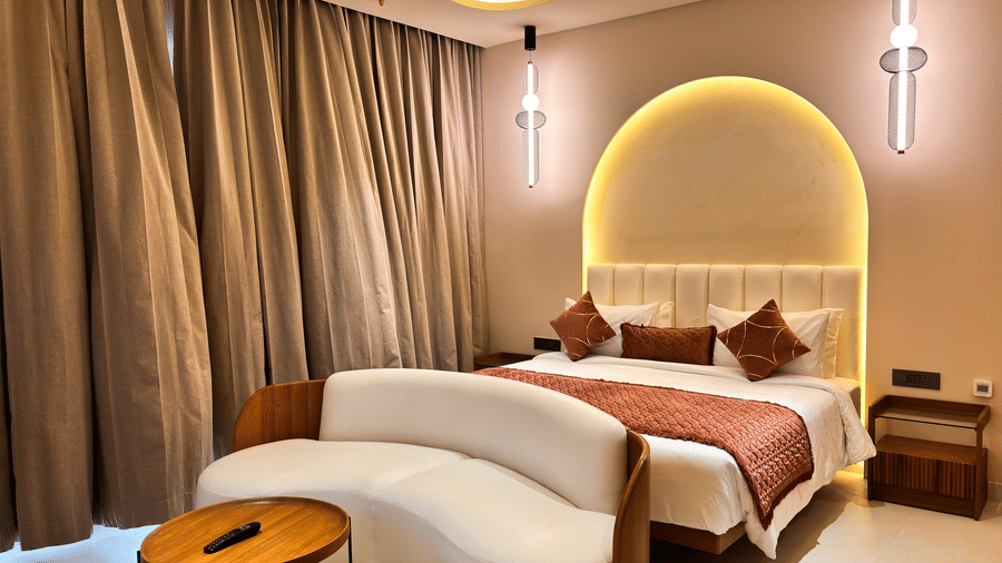 A bed with a brown runner sits behind a white sofa and a wooden table under 2 modern hanging lights at Avanya Resorts N Retreat.