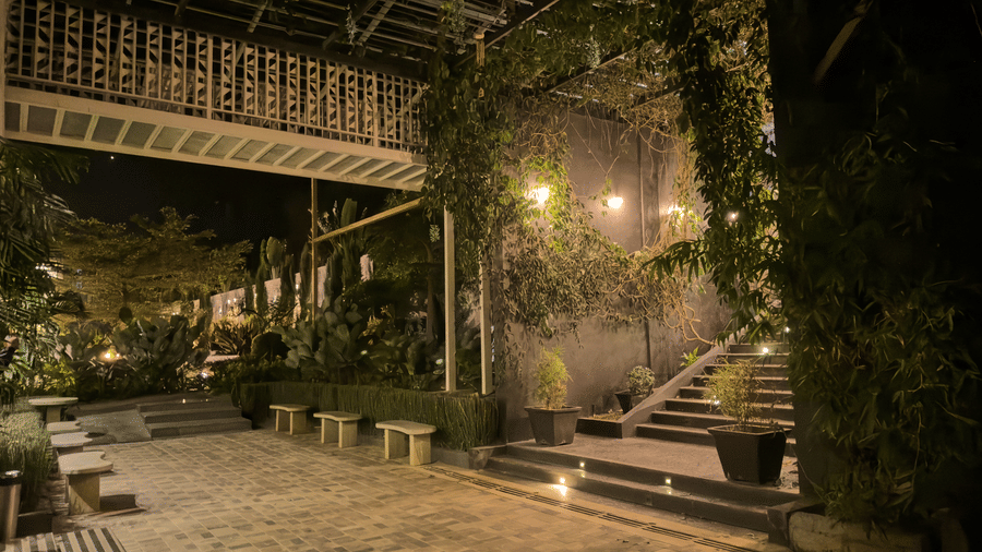 A stone-paved outdoor courtyard area at night with steps, benches, overhead metal structures, and green climbing plants at Bindiga Peak Resort.