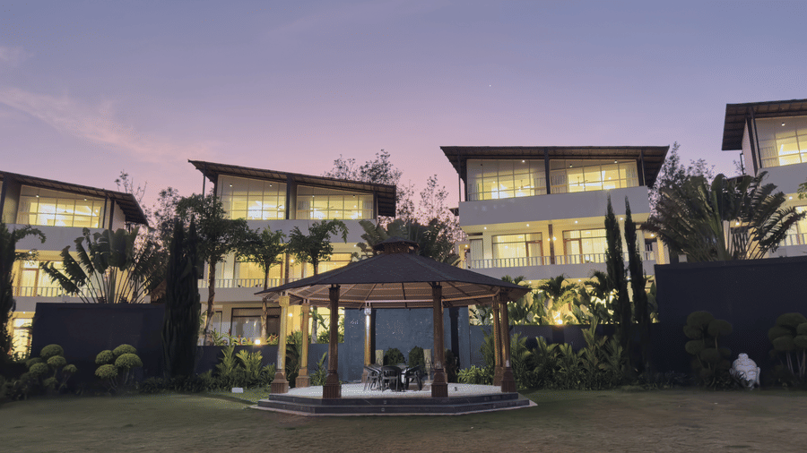 A wooden gazebo on a lawn in front of multi-storey white villas with lights on during dusk at Bindiga Peak Resort.