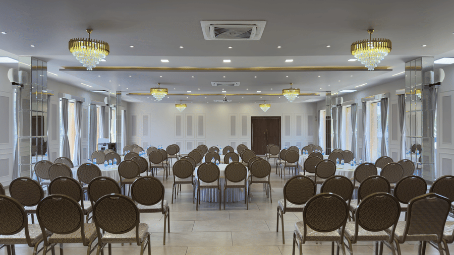 A spacious indoor banquet hall featuring rows of dark grey chairs, white walls, tiled flooring, and hanging chandeliers at Bindiga Peak Resort.