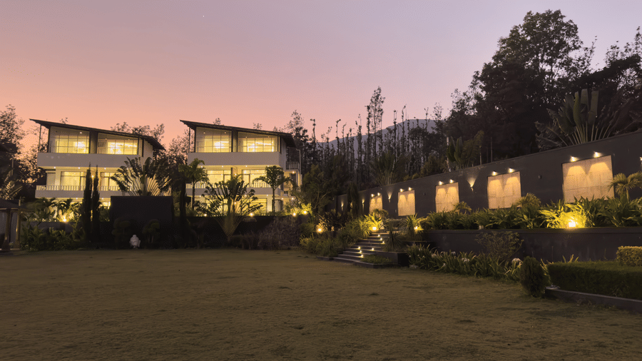 Multi-storey white villas and a lit stone wall bordering a large open lawn under a pink evening sky at Bindiga Peak Resort.