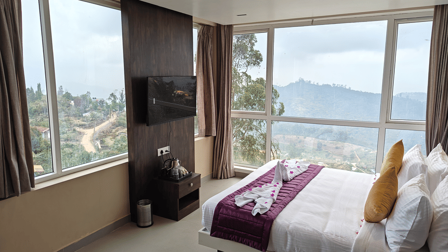 A bedroom view from a high angle showing a double bed and a television mounted on a dark wood wall at Bliss Casa Mila By Bergamont Hotels, Kodaikanal.