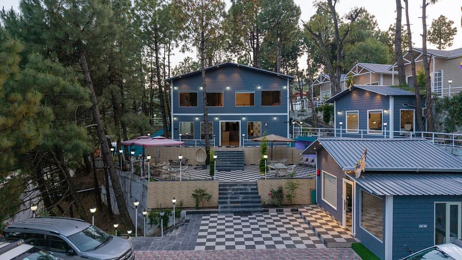 Two modern buildings nestled among tall pine trees with parked cars in the foreground - DLS Nature Trinket Resort, Dalhousie.