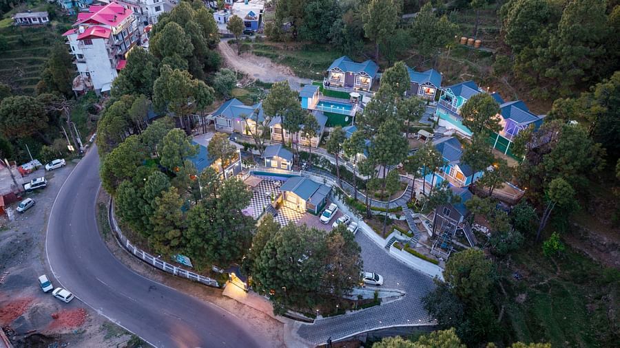 Aerial view of a hillside resort complex with blue-roofed cottages, located alongside a winding road and surrounded by lush greenery and nearby town buildings - DLS Nature Trinket Resort, Dalhousie.