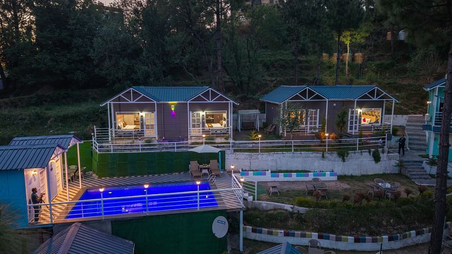 Evening view of a hillside resort with small, well-lit cottages and a glowing blue swimming pool, surrounded by trees and nestled in a peaceful forested area - DLS Nature Trinket Resort, Dalhousie.