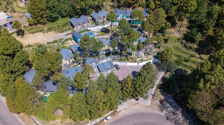 Aerial view of multiple buildings nestled in a lush green, tree-filled landscape next to a winding road - DLS Nature Trinket Resort, Dalhousie.
