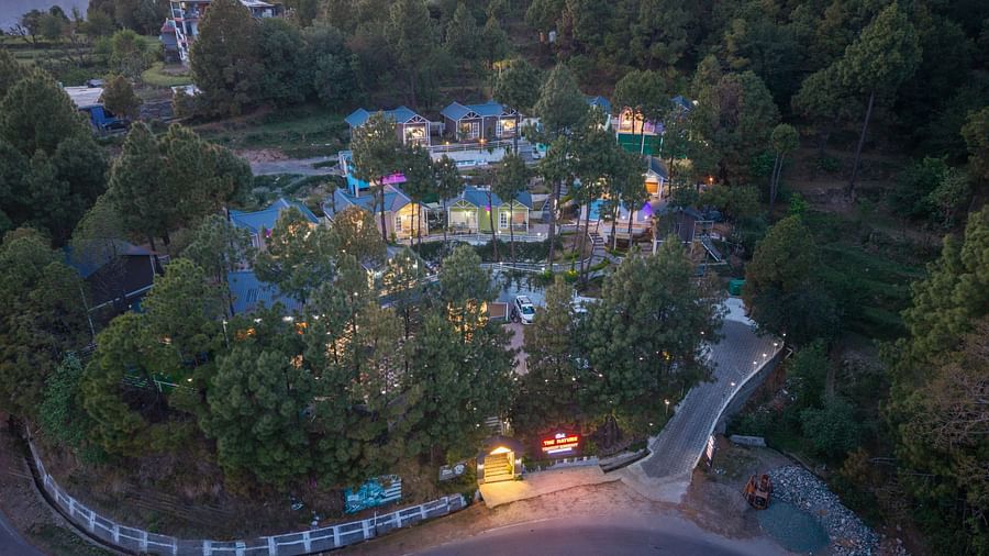 An aerial view shows a hillside resort with multiple buildings nestled among trees at dusk - DLS Nature Trinket Resort, Dalhousie.