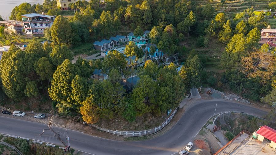 Aerial view of a hillside resort surrounded by dense greenery, located on a sharp bend in a mountain road with scattered buildings and terraced slopes in the background - DLS Nature Trinket Resort, Dalhousie.