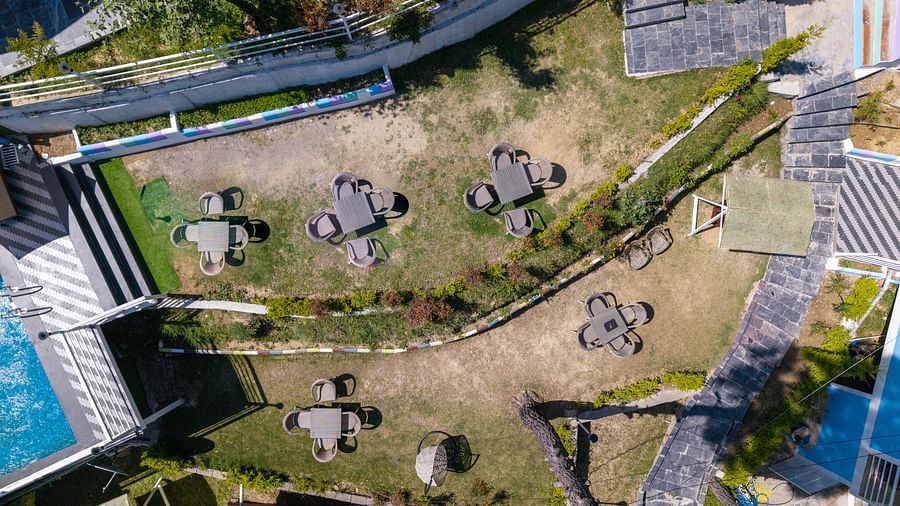 Aerial view of a terraced garden area with multiple round tables and chairs arranged on grassy levels, surrounded by walkways, plants, and nearby buildings - DLS Nature Trinket Resort, Dalhousie.
