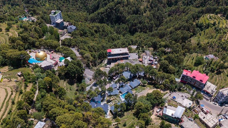 Aerial view of a complex of buildings nestled on a hillside covered with pine trees, with a golden dome visible - DLS Nature Trinket Resort, Dalhousie.