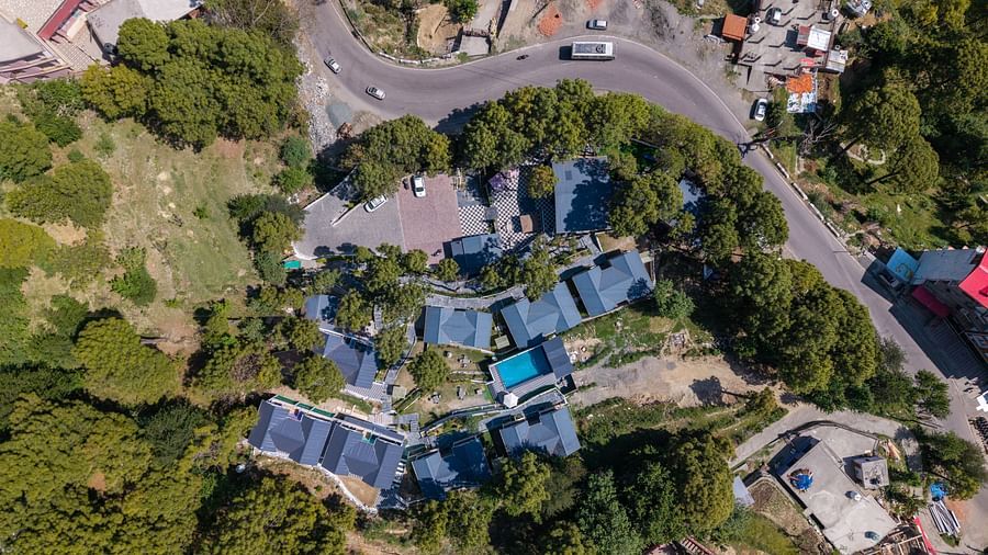 Top-down shot of blue-roofed buildings among dense trees, beside a curved road with a white vehicle - DLS Nature Trinket Resort, Dalhousie.