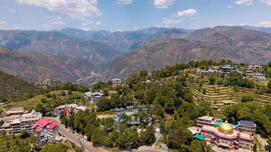 An aerial view of rolling hills, with residential and commercial buildings among lush greenery. A winding road connects the structures, and a temple with a golden dome stands out - DLS Nature Trinket Resort, Dalhousie.