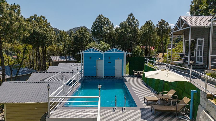 Elevated view of a rectangular swimming pool surrounded by lounge chairs, umbrellas, and colourful cabins, set amidst tall pine trees and hills under a clear sky - DLS Nature Trinket Resort, Dalhousie.