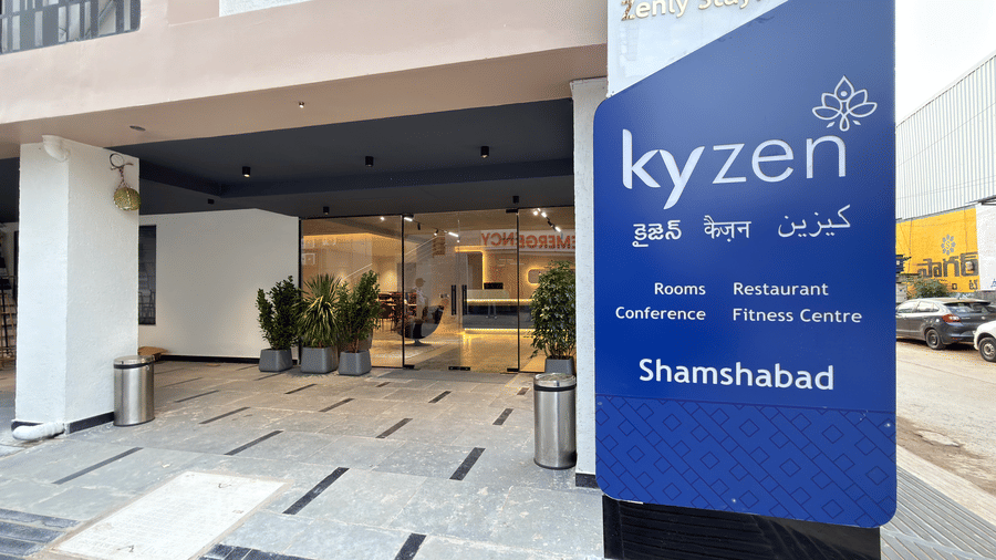 Kyzen hotel entrance at Shamshabad featuring glass doors, landscaped planters, a welcoming signboard, and a security guard at the entrance.