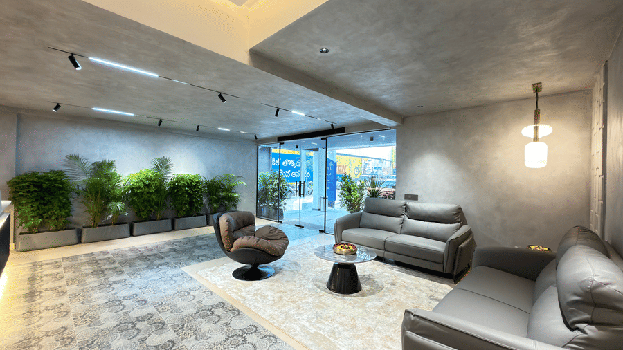 Contemporary hotel lobby lounge with grey sofas, a statement armchair, indoor plants, patterned flooring, and glass entrance doors.