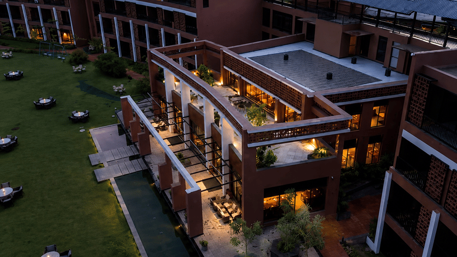 An elevated view at dusk of Hotel Sonar Bangla, Susunia featuring illuminated terrace seating, brick building facades, and lawn tables.