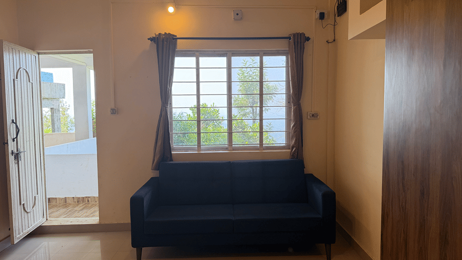 A dark blue sofa placed beneath a window with brown curtains in a neutrally painted room | Kollanoor Guest House, Coorg