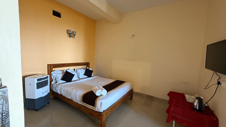 A bedroom with light yellow walls featuring a wooden double bed, an air cooler, and a wall-mounted television | Kollanoor Guest House, Coorg