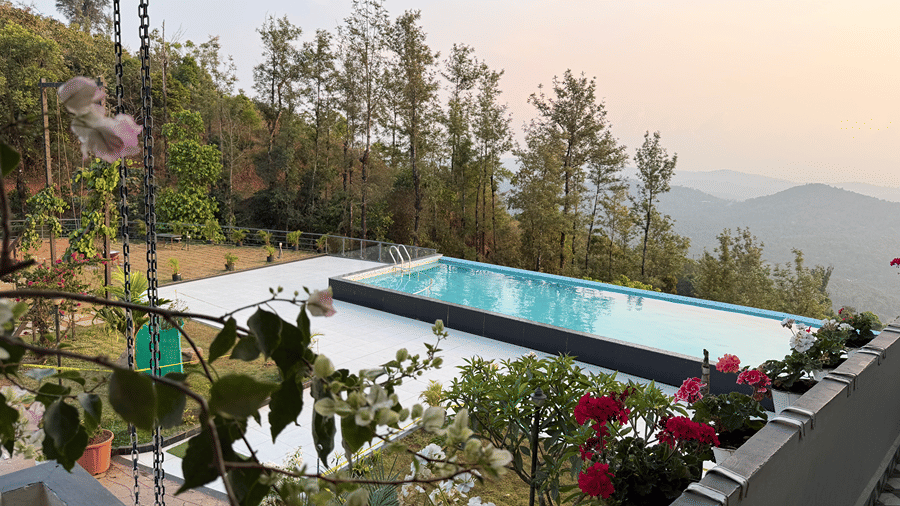 An outdoor swimming pool surrounded by a paved deck, potted flowers, and green trees with hills in the background | Kollanoor Guest House, Coorg
