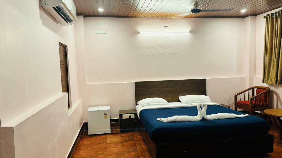 A wide angle view of a bedroom with a queen bed, a mini fridge,  a wooden table and chairs, and green curtains at Paradise Lagoon Resort, Udupi.