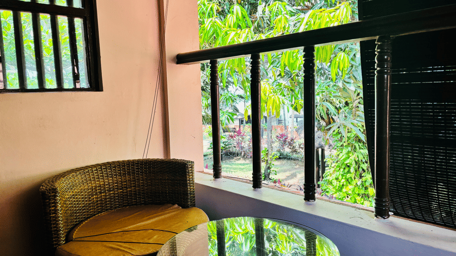 Balcony with round glass-top table and a wicker chair overlooking greenery at Paradise Lagoon Resort, Udupi.