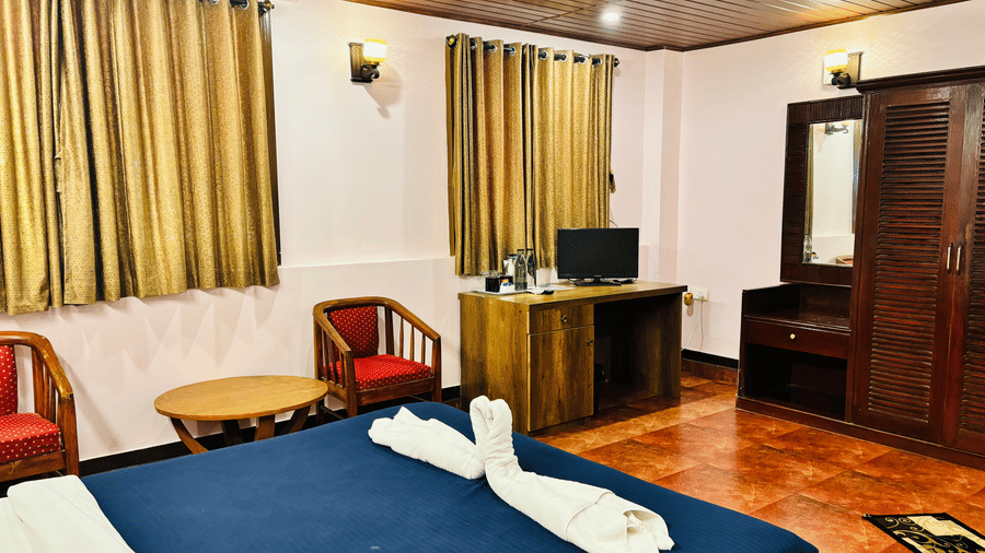 Interior of a standard room at Paradise Lagoon Resort, Udupi, featuring a double bed, wooden furniture including a desk and wardrobe, gold-colored curtains, and a seating area with red cushions.
