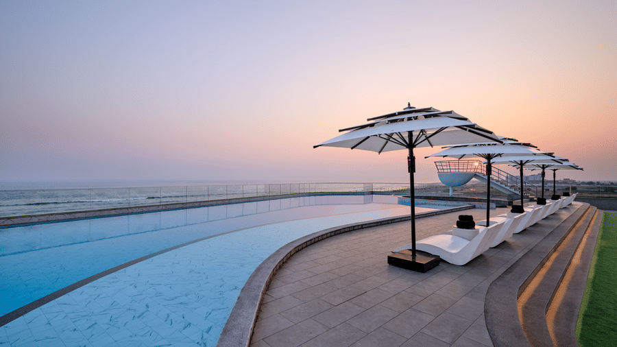 The pool at Swosti Premium Beach Resorts, Puri, with several lounge chairs and white umbrellas lined up along the curved edge of the pool.