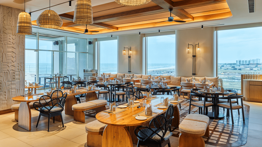 The well-lit Beach Hut at Swosti Premium Beach Resorts, Puri, featuring wooden accents, multiple dining tables, and large windows. 