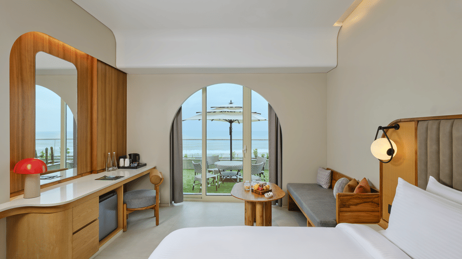 The Premium Room at Swosti Premium Beach Resorts, Puri, featuring a double bed, a television mounted on the wall, modern wooden decor, and large windows displaying a  sea view. 