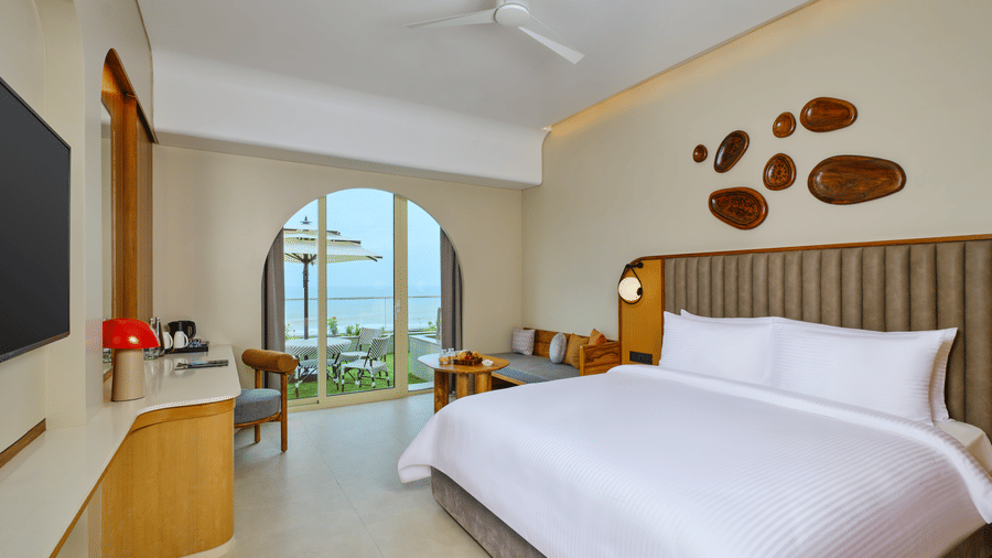 The Premium Room at Swosti Premium Beach Resorts, Puri, with a double bed, a television mounted on the wall, modern wooden decor, and large windows displaying a  sea view. 