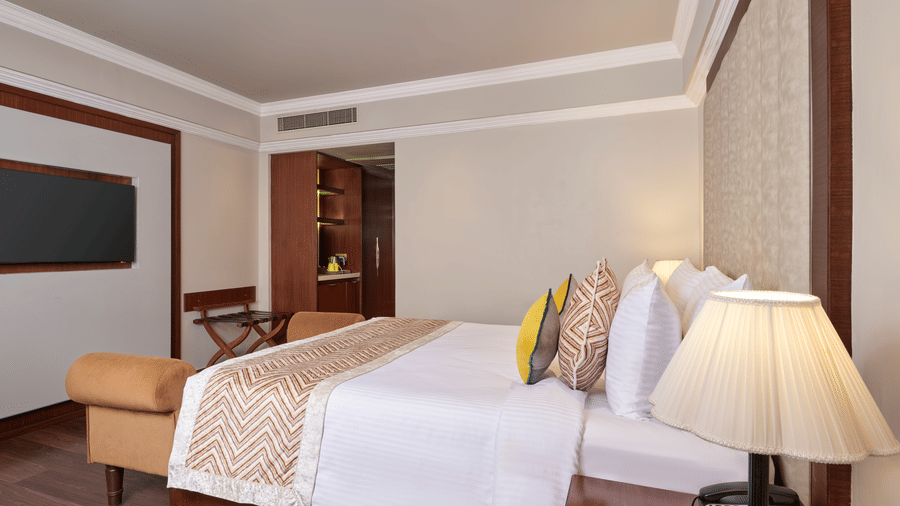 The Club Room at Swosti Premium, Bhubaneswar, featuring double bed, bedspreads, pillows,  a lamp, wooden tables, a cupboard, and a television.