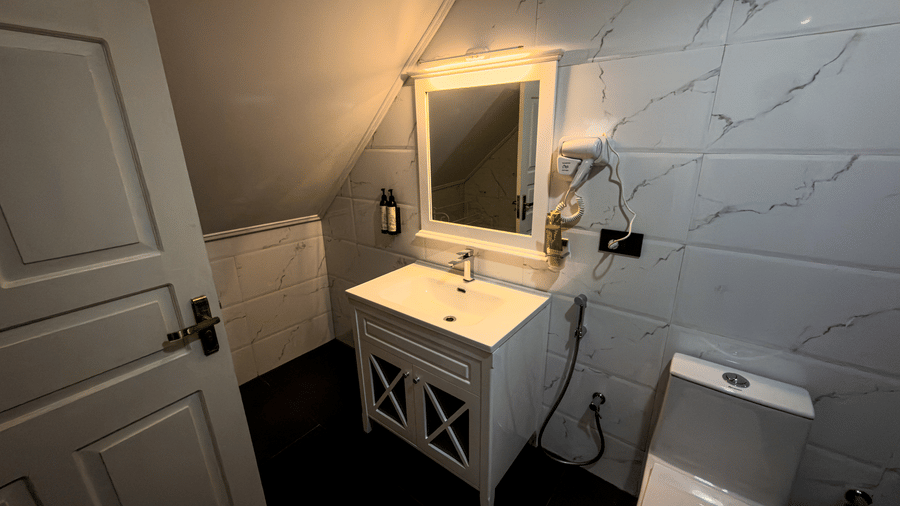 The vanity area in the bathroom of the Tagore Suite at The Heritage Club Tripura Castle Hotel and Spa, Shillong, featuring a bright mirror and a simple sink under a sloped wall.