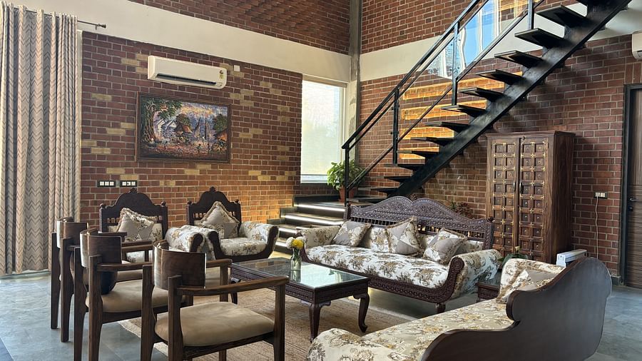 A living room at Tisya Farms, Gurgaon with multiple sofas, large windows, brick walls, and an overhead staircase.
