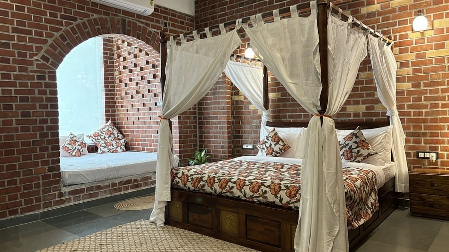 A bedroom at Tisya Farms, Gurgaon with a four-poster bed, white curtains, brick walls, and an arched window.