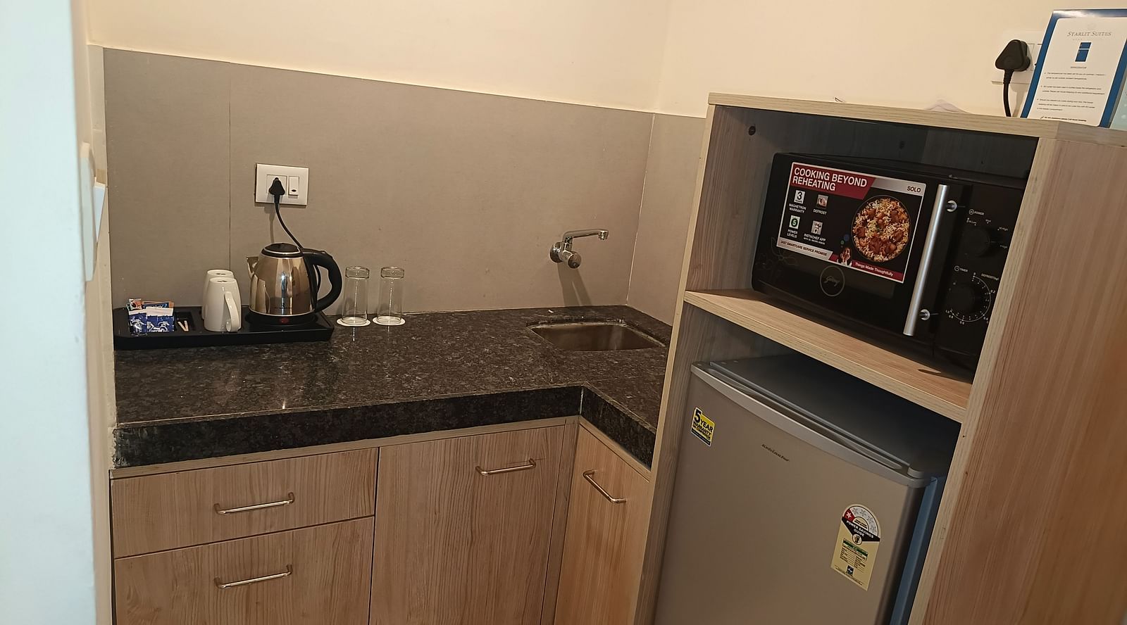 Kitchenette inside Premium Studio at Starlit Suites, Shirdi features light wood cabinets, a dark countertop with a kettle and cups, a small sink, a microwave, and a mini-refrigerator unit.