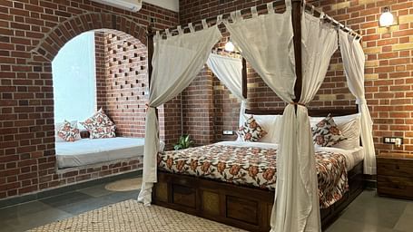 A bedroom at Tisya Farms, Gurgaon with a four-poster bed, white curtains, brick walls, and an arched window.