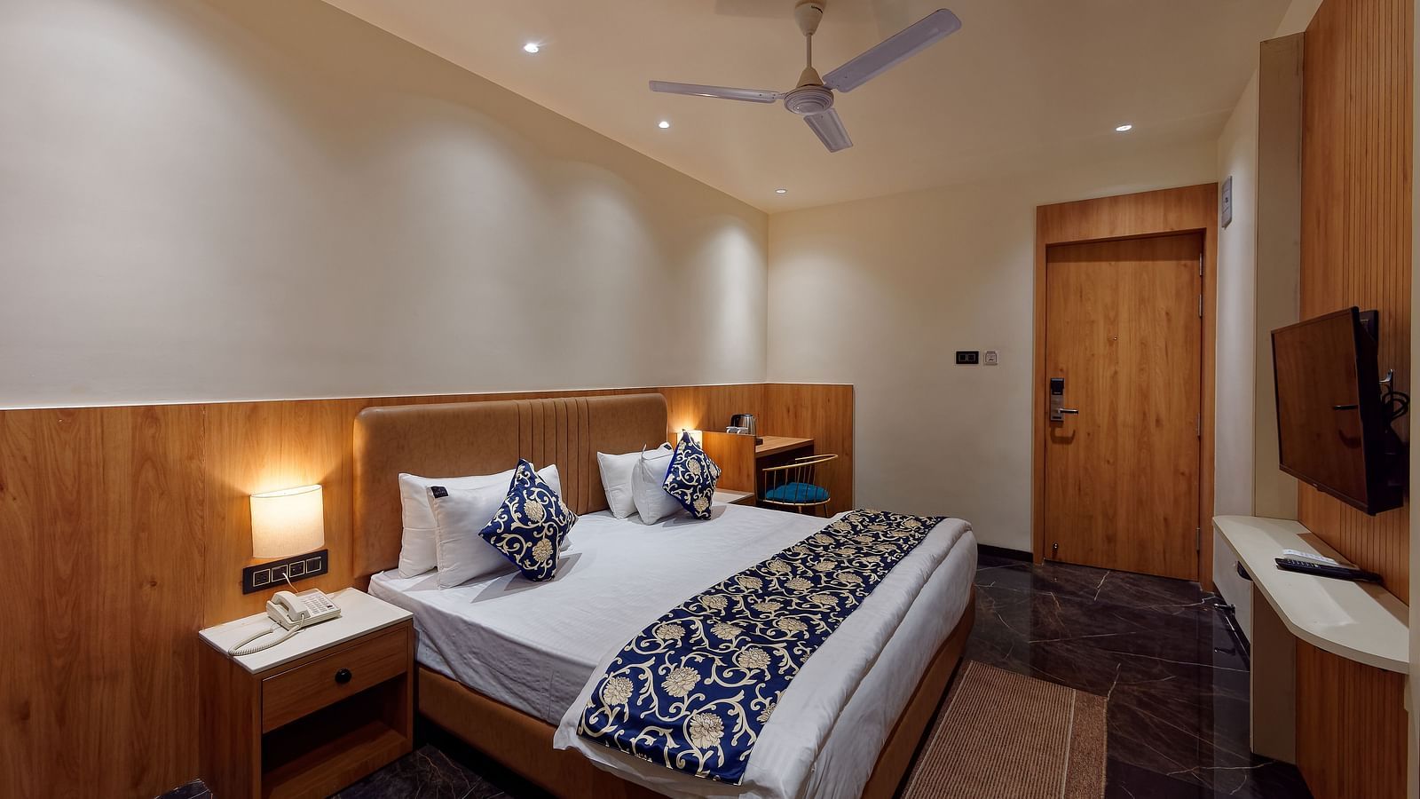 Image of a studio room with a bed with a table on the side of the bed and a television in front Image of a bed with table and chair on the side of the table and a fan on te ceiling at Ankit Hotel, Jabalpur