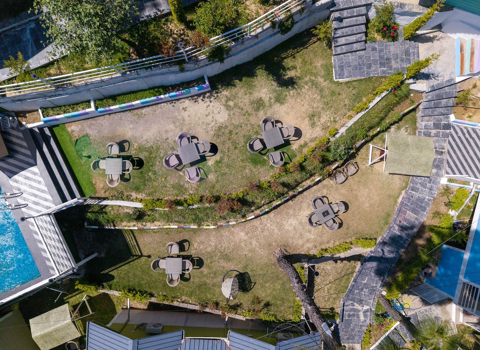 Aerial view of a terraced garden area with multiple round tables and chairs arranged on grassy levels, surrounded by walkways, plants, and nearby buildings - DLS Nature Trinket Resort, Dalhousie.