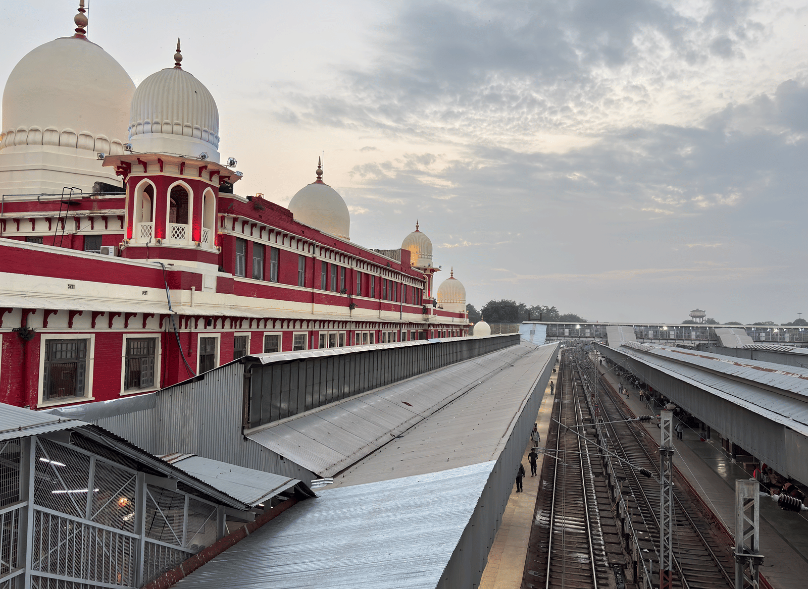 Varanasi Junction railway station with its red and white building topped by white domes, metal platform roofs, and railway tracks under a cloudy sky.
