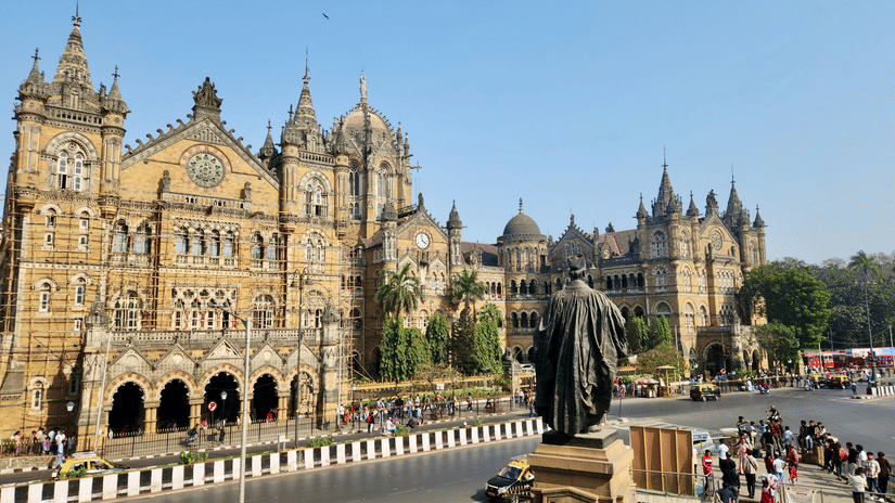Chhatrapati Shivaji Maharaj Terminus in Mumbai with its historic Victorian Gothic architecture, a statue in the foreground, and a busy road with vehicles and pedestrians under a clear sky.