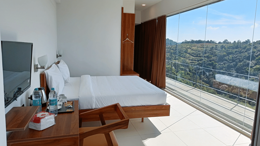 Second Floor Corner Room at The Cliff Edge Coorg Hotel featuring a bed, TV, and a work desk, with panoramic forest views visible through massive floor-to-ceiling windows.